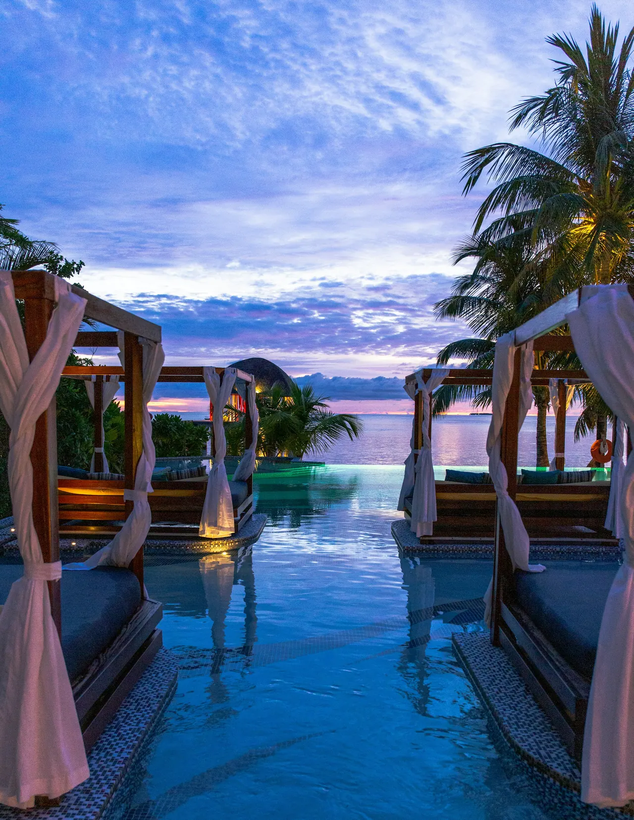 Poolside cabanas with draped curtains at dusk, the sea beyond